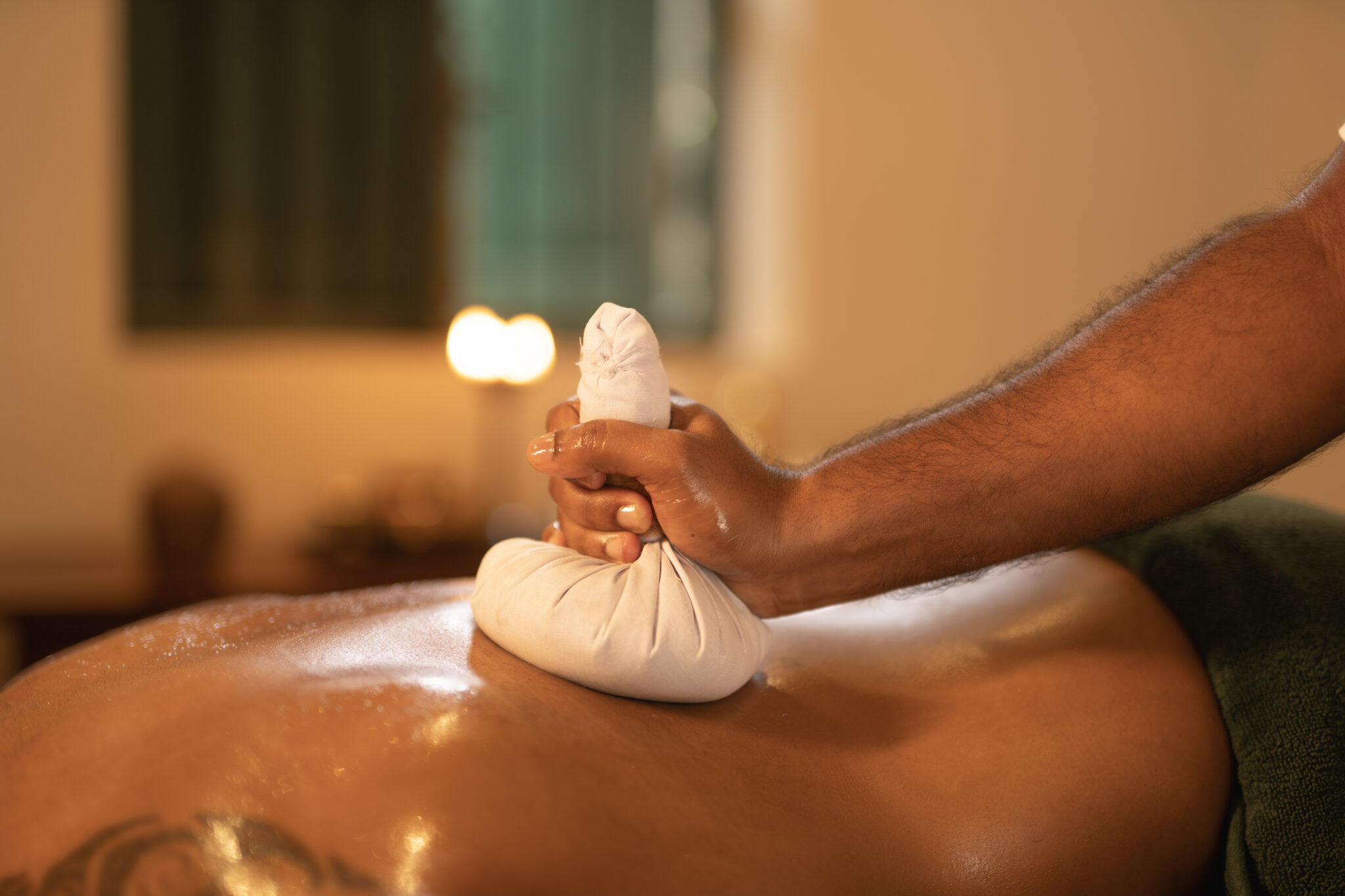 In a calm, softly lit room, a person receives a warm herbal compress massage on their back, offering a serene cleanse to help detox the body and mind. Resplendent Ceylon